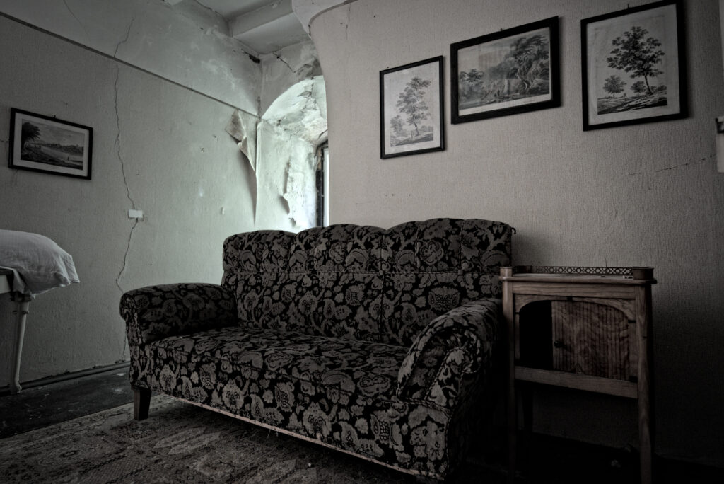 Ein Sofa, in einem Heruntergekommenen Raum. An der Wand hängen drei Bilder mit Bäumen darauf. Einige Risse in der Wand