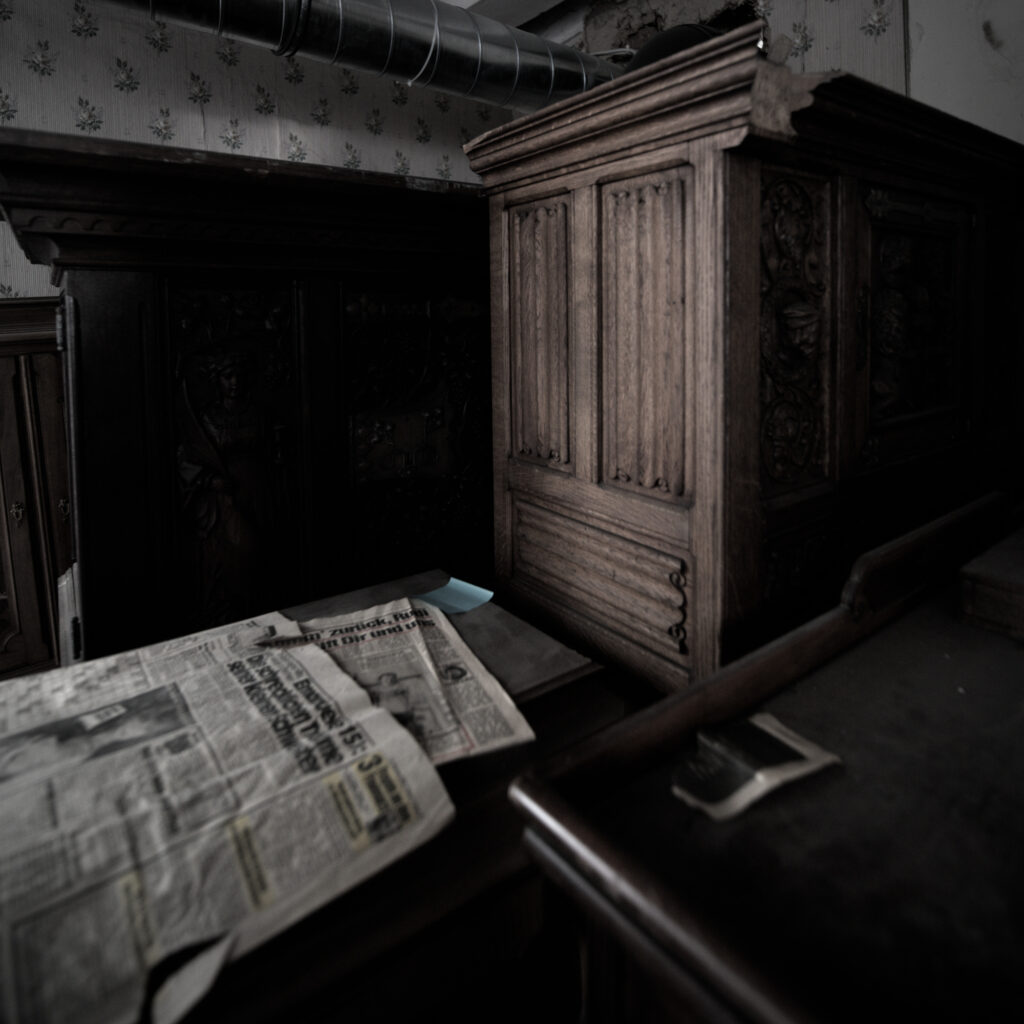 Ein unaufgeräumter Raum. Darin steht ein Holzschrank und davor zwei Holztische. Auf einem der Tische liegen einzelne Seiten einer alten Zeitung
