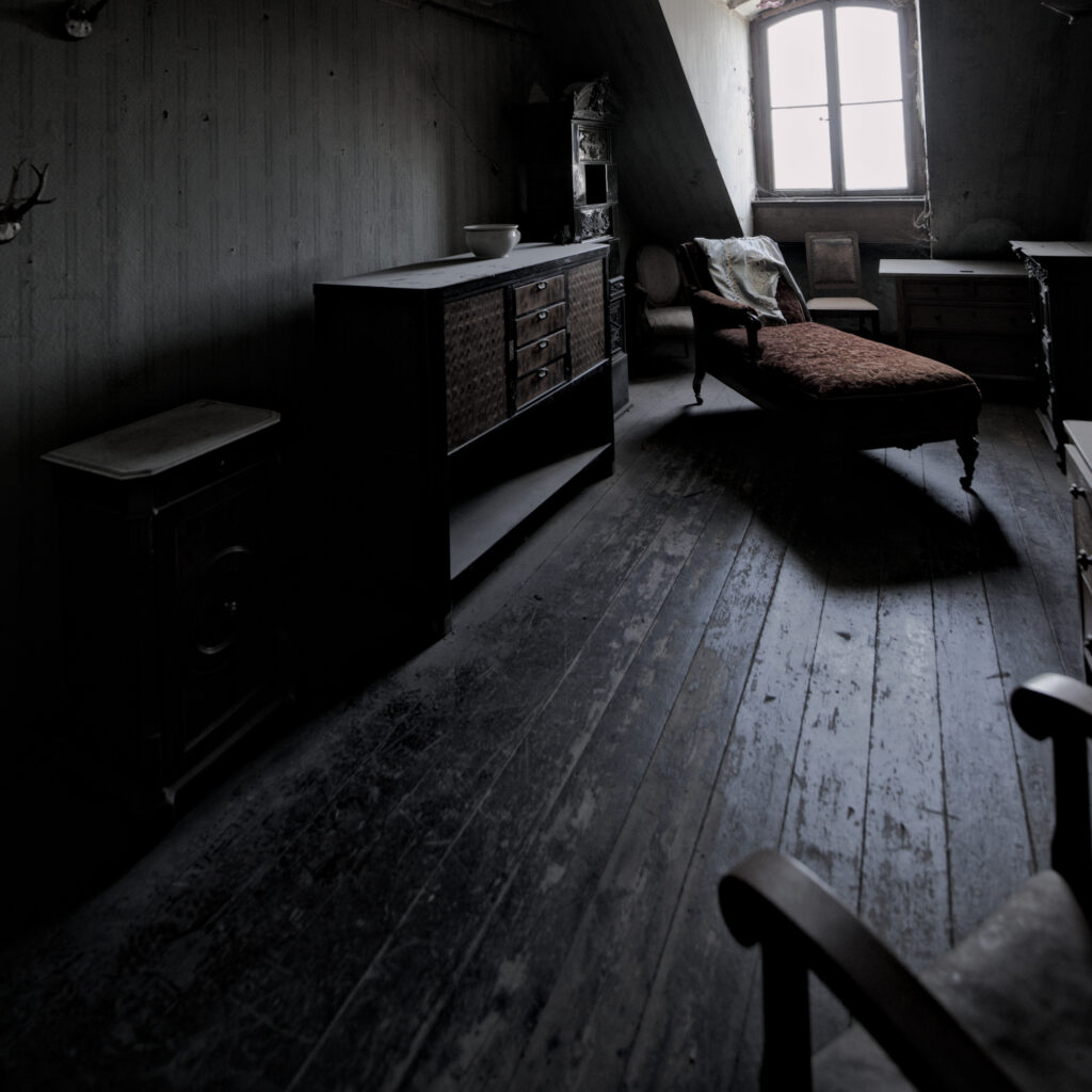 Auf einem Dachboden. Links an der Wand steht eine alte Kommode. Rechts daneben eine Reacmere