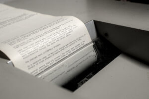 Detailaufnahme von einem Siemens Fernschreiber. Es erinnert ein wenig an ein Faxgerät. Es hängt ein beschriftetes Papier aus dem Gerät heraus.