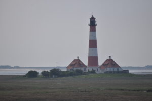 Ein rot-weißer Leuchtturm mit einem roten Licht an der Spitze steht auf einer kleinen Insel. Im Vordergrund sind grüne Wiesen mit Schafen zu sehen.