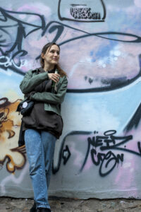 Eine Person lehnt an einer Wand mit Graffiti, trägt eine grüne Jacke und eine braune Kapuzenjacke, hält ein Getränk in der Hand. Neben ihr steht eine weitere Person in Lederjacke und kariertem Hemd.