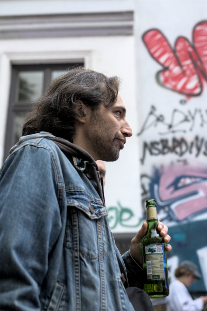 Junger Mann mit langen, lockigen Haaren trägt eine Jeansjacke und hält eine grüne Flasche in der rechten Hand. Im Hintergrund ist ein Graffiti mit roten und pinken Elementen zu sehen.