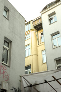 Blick in einen Innenhof zwischen zwei weißen Gebäuden. Ein gelbes Gebäude mit Balkonen ist im Hintergrund sichtbar. Ein Teil eines weißen Zeltes ist im Vordergrund.