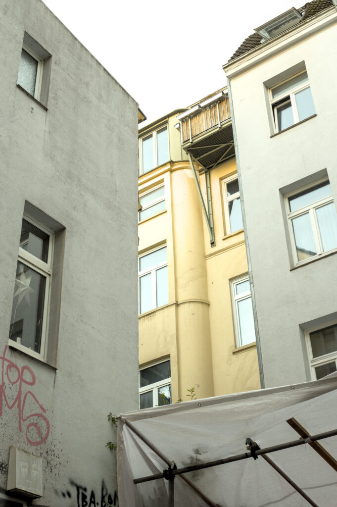 Blick in einen Innenhof zwischen zwei weißen Gebäuden. Ein gelbes Gebäude mit Balkonen ist im Hintergrund sichtbar. Ein Teil eines weißen Zeltes ist im Vordergrund.