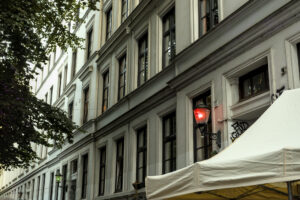 Fassade eines mehrstöckigen Gebäudes mit grauer Wandfarbe und mehreren Fenstern. Ein rotes Licht leuchtet an einer Wandlampe. Im Vordergrund steht ein weißes Zelt.