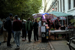 Eine Gruppe von Menschen steht in einer engen Gasse vor einem Stand mit bunten Plakaten und einem grünen Pavillon. Im Hintergrund sind Bäume und ein Gebäude sichtbar.