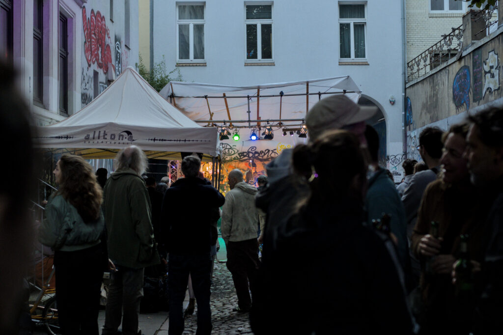 Menschenmenge in einer Gasse, die auf eine Bühne mit bunten Lichtern schaut. Ein Zelt mit der Aufschrift ‘alltona’ steht im Vordergrund.