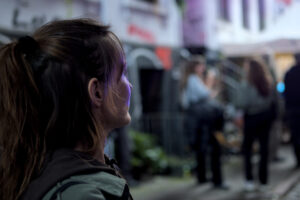 Eine Person mit langen, braunen Haaren und einem Pferdeschwanz steht im Vordergrund. Im Hintergrund sind mehrere Personen in einer urbanen Umgebung zu sehen.