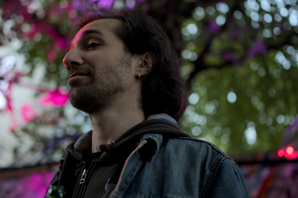 Ein Mann mit langen, dunklen Haaren trägt eine graue Kapuzenjacke und eine Jeansjacke. Im Hintergrund sind grüne Blätter und bunte Lichter sichtbar.