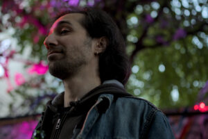 Ein Mann mit langen, dunklen Haaren trägt eine graue Kapuzenjacke und eine Jeansjacke. Im Hintergrund sind grüne Blätter und bunte Lichter sichtbar.