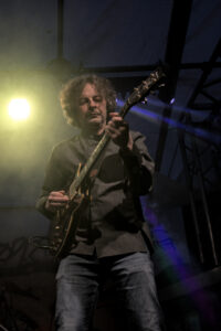 Ein Musiker spielt eine E-Gitarre auf einer Bühne. Er trägt ein graues Hemd und Jeans. Scheinwerferlicht strahlt auf ihn und erzeugt farbige Lichtstrahlen.