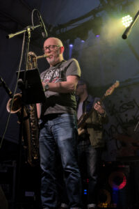Ein Musiker spielt Saxophon auf einer Bühne, trägt ein graues T-Shirt und Jeans. Im Hintergrund steht ein weiterer Musiker mit einer E-Bass.