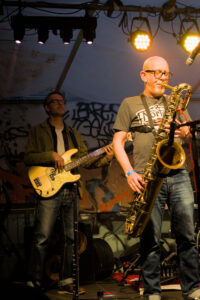 Ein Musiker spielt Saxophon auf einer Bühne, trägt ein graues T-Shirt und Jeans. Im Hintergrund steht ein weiterer Musiker mit einer E-Bass.