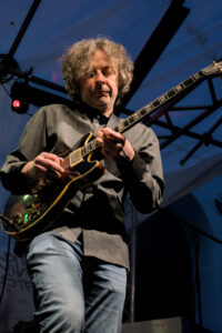 Ein Musiker spielt eine E-Gitarre auf einer Bühne. Er trägt ein graues Hemd und Jeans. Scheinwerferlicht strahlt auf ihn und erzeugt farbige Lichtstrahlen.