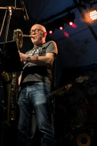 Ein Musiker spielt Saxophon auf einer Bühne, trägt ein graues T-Shirt und Jeans. Im Hintergrund steht ein weiterer Musiker mit einer E-Bass.