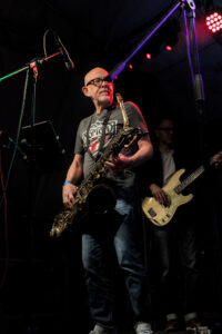 Ein Musiker spielt Saxophon auf einer Bühne, trägt ein graues T-Shirt und Jeans. Im Hintergrund steht ein weiterer Musiker mit einer E-Bass.