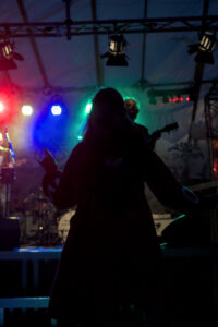 Silhouette einer Person in einem roten Mantel, die mit einer Hand ein Peace-Zeichen macht. Bunte Lichter in grün, blau und rot leuchten im Hintergrund.