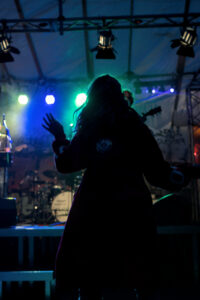 Silhouette einer Person in einem roten Mantel, die mit einer Hand ein Peace-Zeichen macht. Bunte Lichter in grün, blau und rot leuchten im Hintergrund.