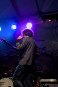 Ein Musiker spielt eine E-Gitarre auf einer Bühne. Er trägt ein graues Hemd und Jeans. Scheinwerferlicht strahlt auf ihn und erzeugt farbige Lichtstrahlen.