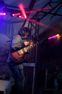 Ein Musiker spielt eine E-Gitarre auf einer Bühne. Er trägt ein graues Hemd und Jeans. Scheinwerferlicht strahlt auf ihn und erzeugt farbige Lichtstrahlen.