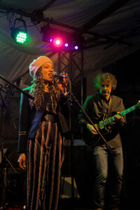 Eine Sängerin mit langen, lockigen Haaren trägt einen weißen Hut und ein gestreiftes Outfit. Sie hält ein Mikrofon und singt auf einer Bühne. Ein Gitarrist im Hintergrund spielt eine schwarze Gitarre.