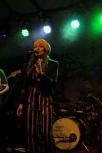 Eine Sängerin mit langen, lockigen Haaren trägt einen weißen Hut und ein gestreiftes Outfit. Sie hält ein Mikrofon und singt auf einer Bühne. Ein Gitarrist im Hintergrund spielt eine schwarze Gitarre.