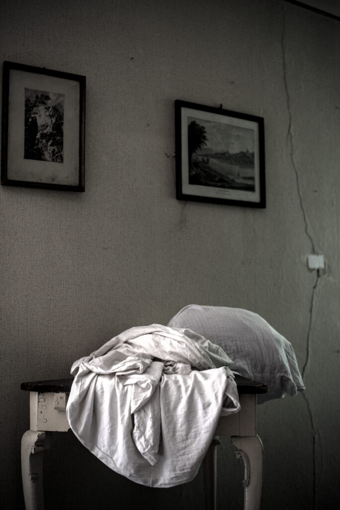 Auf einem Holzitisch liegen eine Bettdecke und ein Kissen. An der Wand dahinter zieht sich ein langer Riss. Daneben hängen zwei Bilder