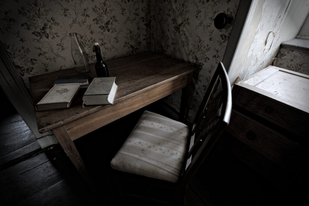 Ein Holztisch. Davor ein Stuhl und auf dem Tisch liegen drei Bücher. Neben den Büchern stehen zwei Flaschen.
