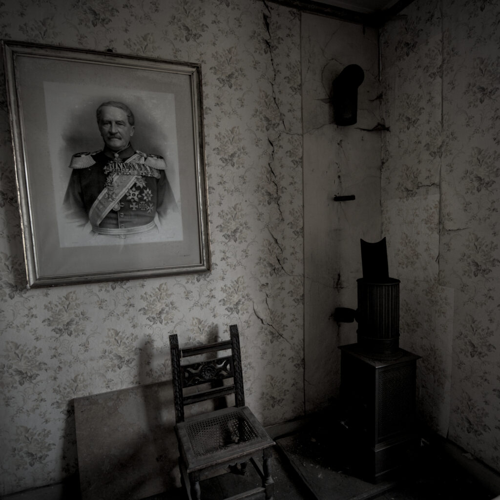 Ein Holzofen in der Ecke eines Raumes. Links daneben hängt an der Wand ein Porträt von einem Mann. Darunter auf dem Boden steht ein Stuhl.