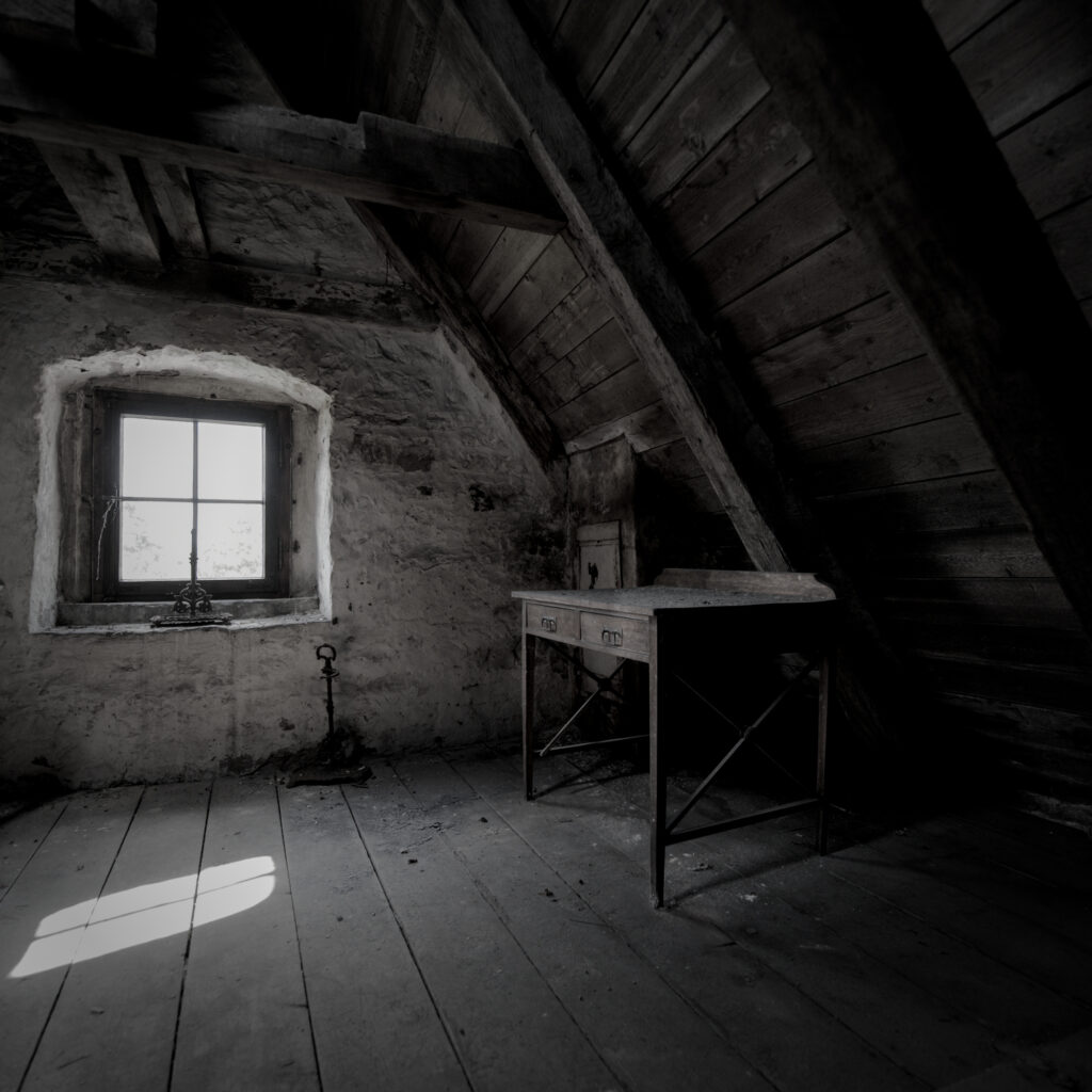 Auf einem Dachboden. An der Wand ein Fenster und unter der Dachschräge ein alter Holztisch