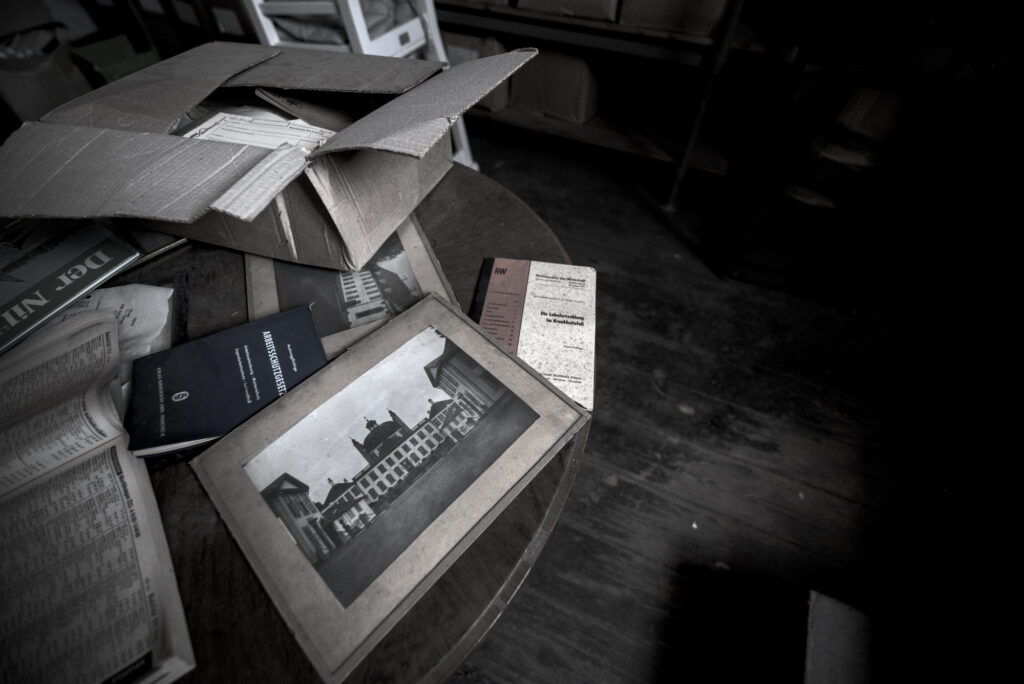Auf einem Runden Holztisch liegen verschiedene Gegenstände. Ein Bild im Rahmen, ein geöffnetes Buch. Vermutlich ein altes Telefonbuch. eine Brouchüre und ein Karton mit weiterem Krempel