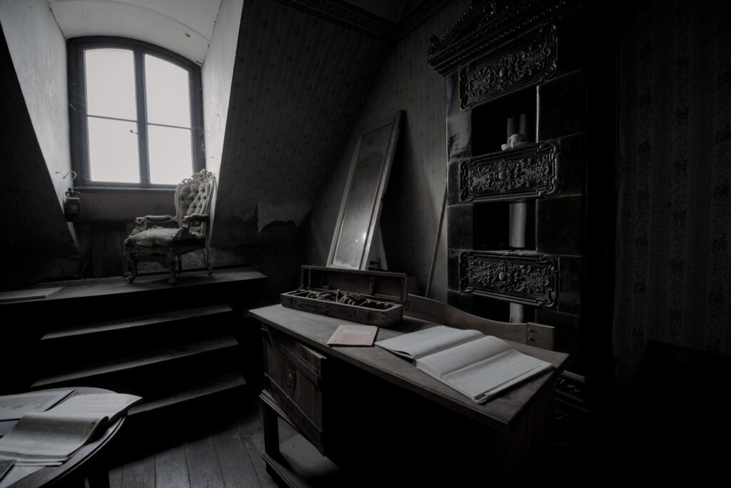 Auf einem Dachboden. An einem Fenster unter einer Dachschräge steht ein alter Sessel auf einer art Podest mit Stufen. Weiter vorne im Bild ein alter Schreibtisch mit einem Geöffneten Buch und einem Holzkasten