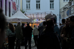Menschenmenge in einer Gasse, die auf eine Bühne mit bunten Lichtern schaut. Ein Zelt mit der Aufschrift 'alltona' steht im Vordergrund.